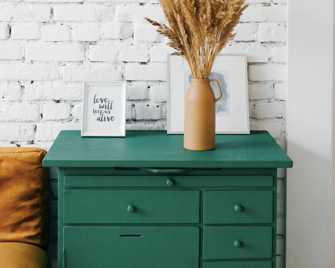 A green dresser holds a mustard vase with dried wheat and framed text reading 'love will keep us alive' against a white brick wall.